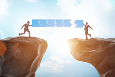 businessman in suits each standing a cliff with a gap between them and holding up puzzle pieces to connect them across the gap