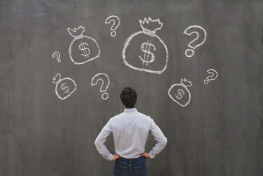 Man staring up at a wall with several drawings on bas with the dollar sign on them and question marks surrounding them
