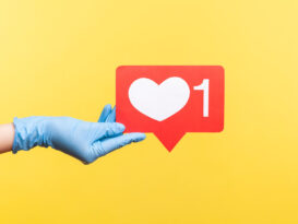 Profile side view closeup of human hand in blue surgical gloves holding social media like stick. indoor, studio shot, isolated on yellow background.