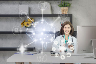 Female doctor sitting at her desk in front of a computer with a image of the outline of a brain in white with various nodes connected around it containing icons representing different digital components including a tablet, email, telecommunications, online shopping, and more