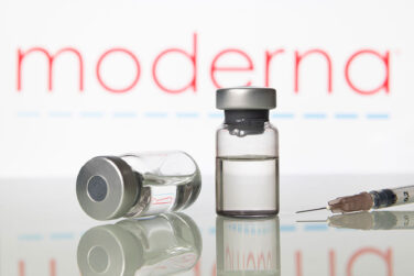 The Moderna logo in the background with vials of a vaccine on a table in front of it